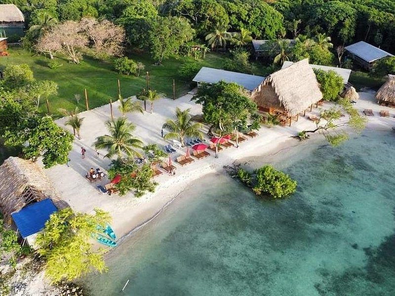 A beach club on a sandy point, thatched roofs and palms over shallow water