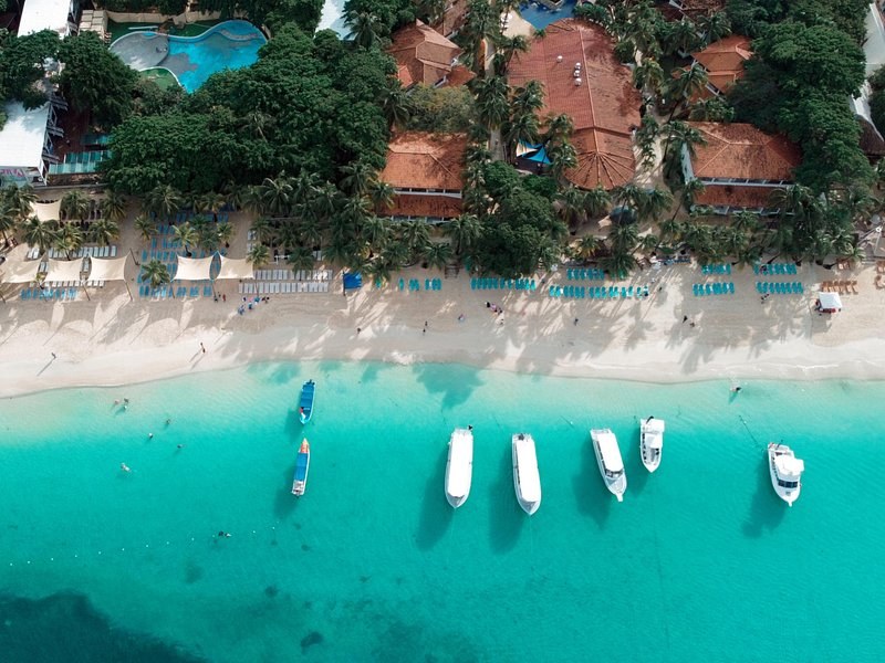 Boats moored in turquoise water off a white beach lined with loungers and palms